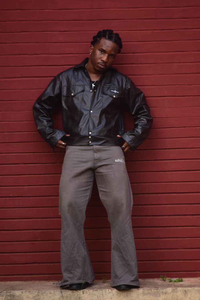 Confident man wearing a leather jacket and flared pants poses outdoors against a red wall.